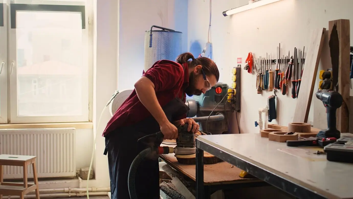 A man wearing protective glasses operates orbital sanding equipment after consulting the schematics.